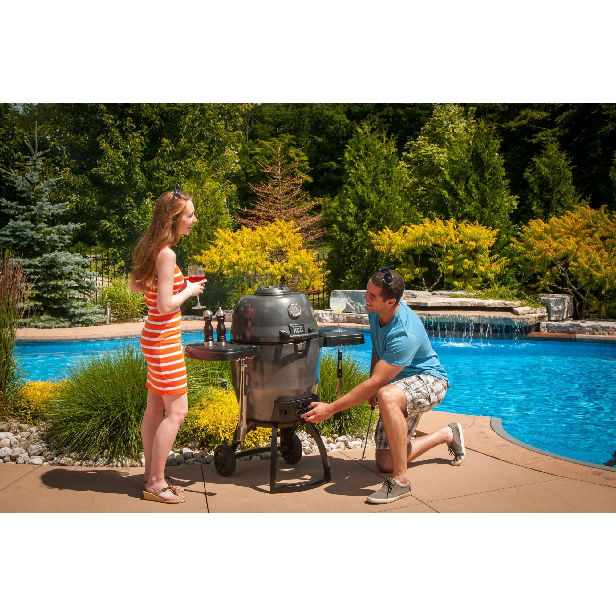 Pareja utilizando la barbacoa Broil King Keg 5000 en una zona de piscina y jardín.