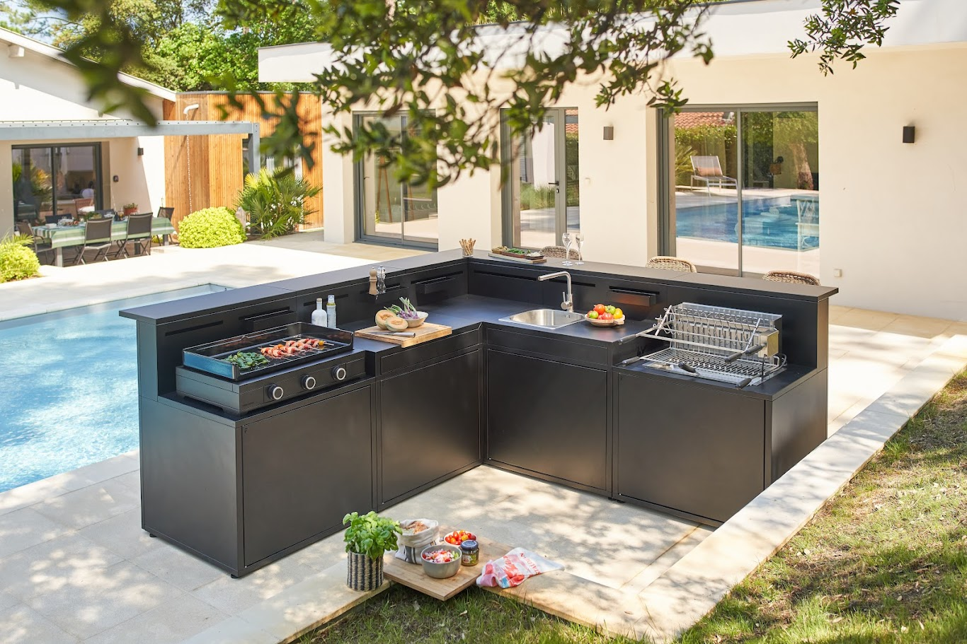 Cocina de exterior modular WMF en configuración de esquina (L) con fregadero, zona de cocción y encimera negra, instalada en un jardín moderno junto a una piscina.