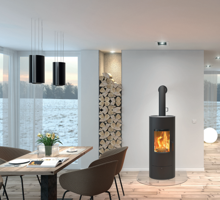 Estufa de pellets Rika Step con diseño cilíndrico negro instalada en un comedor moderno con grandes ventanales y leñero decorativo.